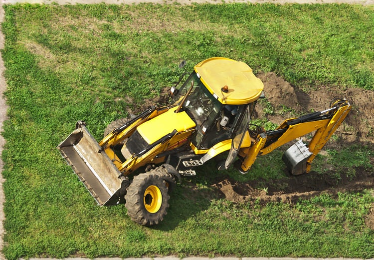 Aerial view of a yellow backhoe digging a trench in a grassy field with a pile of earth beside it.