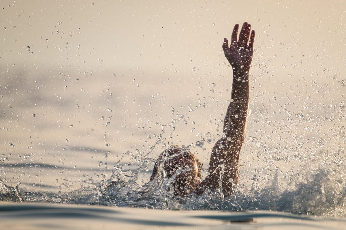 A person’s head and one arm are partially visible above water, surrounded by splashes, with the arm raised.