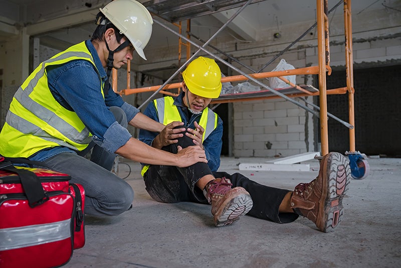 A construction worker, wearing a hard hat and safety vest, kneels to comfort an injured worker sitting on the floor.