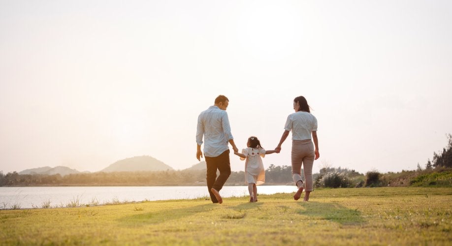 Family of three walking away from the camera, holding hands by a lake with mountains in the distance.
