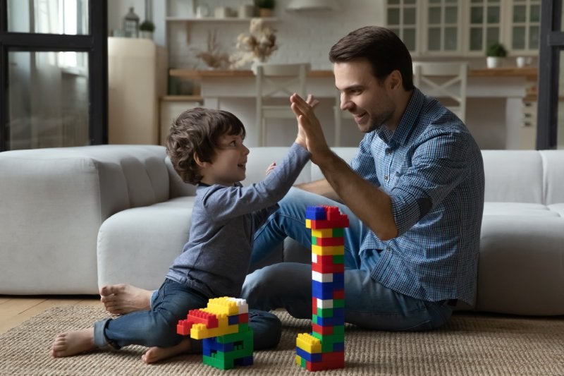 Father and son, sitting on the floor with building blocks, high-fiving in a cheerful living room.