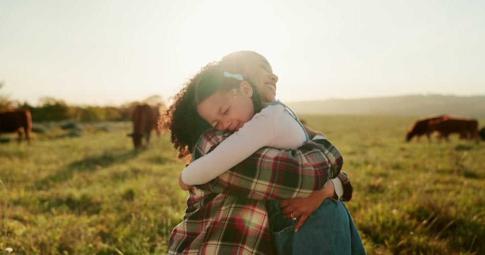 A smiling mother and daughter hugging in a sunny field with cattle grazing in the background.