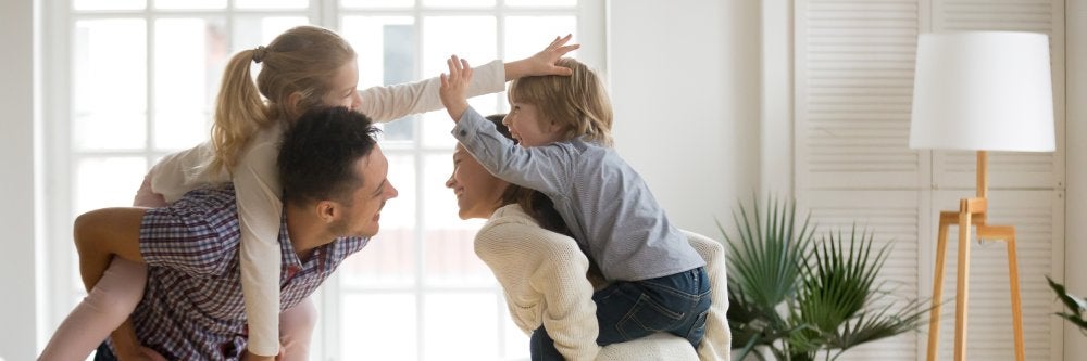 Parents playfully giving their young children piggyback rides indoors.