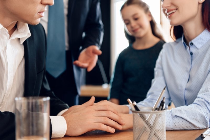 A couple with their hands clasped on a table, a child smiling behind them, and an adult gesturing in the background.