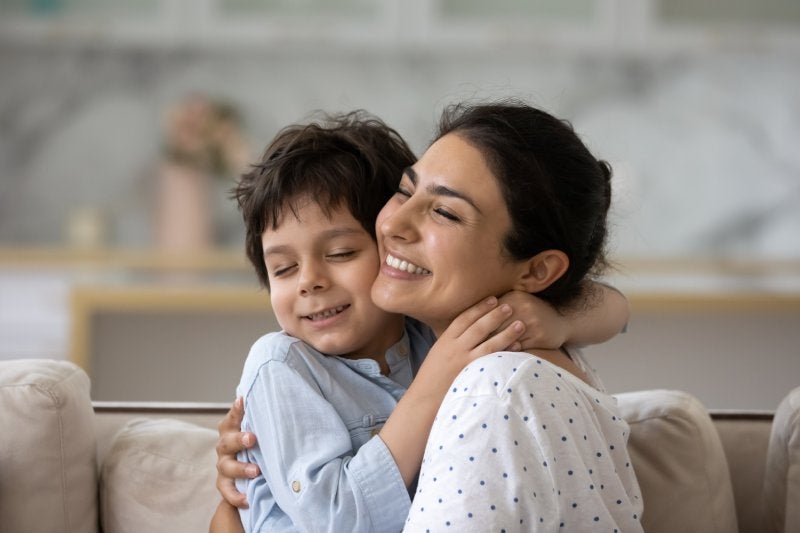 A joyful brown-skinned mother and young son, with closed eyes and big smiles, embracing.