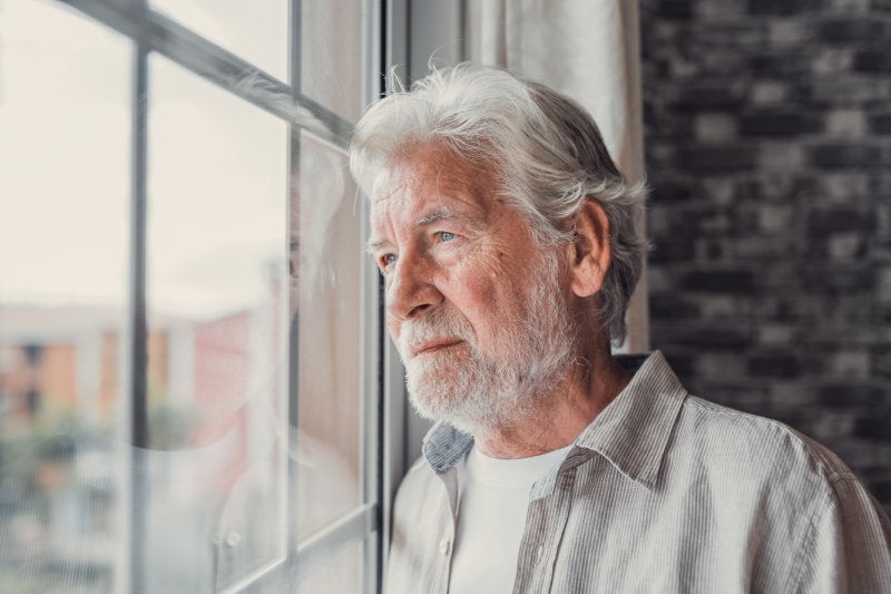 An older man with gray hair and beard sadly looking out a window.