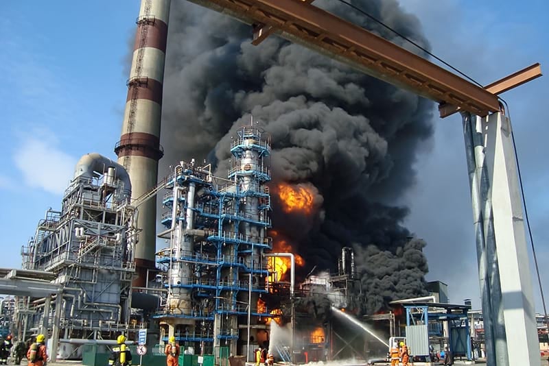 A massive fire at a chemical plant, with black smoke engulfing the structures and firefighters battling the blaze.