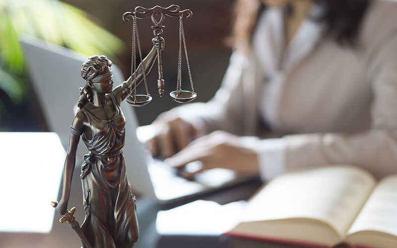 Bronze Lady Justice statue with a blurred person typing on a laptop and an open book in the background.