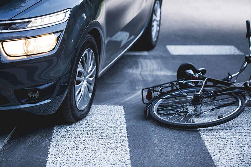 A car with headlights on, parked next to a bicycle lying on its side in a crosswalk after an accident.