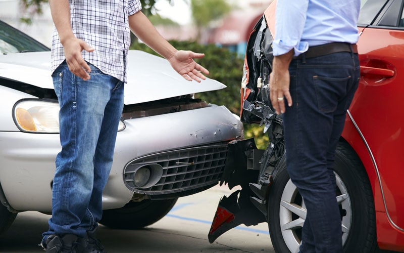 Two people appear to be discussing a damaged silver car and a red car after a collision.