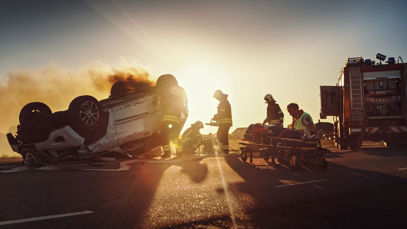 Emergency crew with a flipped, smoking car, tending to a victim near a fire truck at sunset.
