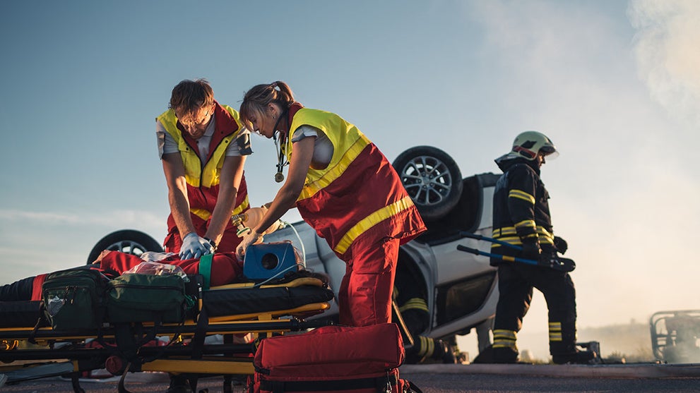 Paramedics performing CPR on a patient on a stretcher beside a flipped car, with a firefighter nearby.