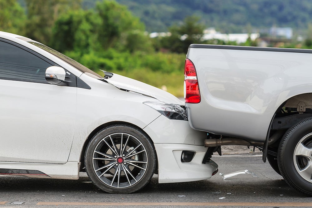 Front of a white car wedged under the back of a silver pickup truck, after a rear-end collision.