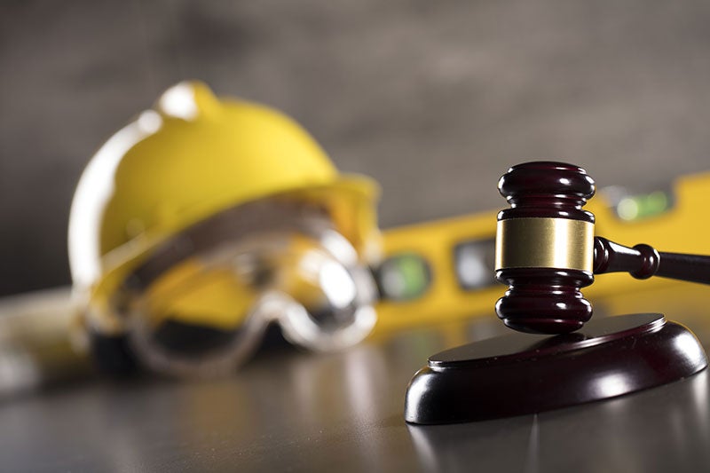 A wooden gavel in the foreground, with a blurry yellow hard hat, safety glasses, and a level in the background.