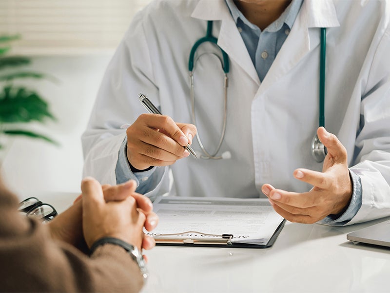 A doctor with a stethoscope writes on a clipboard while speaking with a patient.
