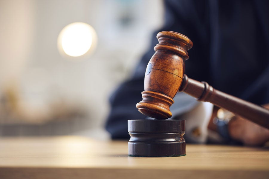 A wooden gavel rests on a round black sound block on a wooden table, with a blurred figure in the background.