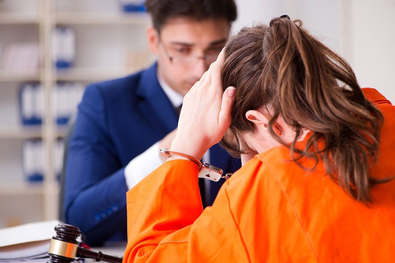 A woman in an orange jumpsuit with handcuffs rubs her forehead while a man in a suit looks on across a desk.