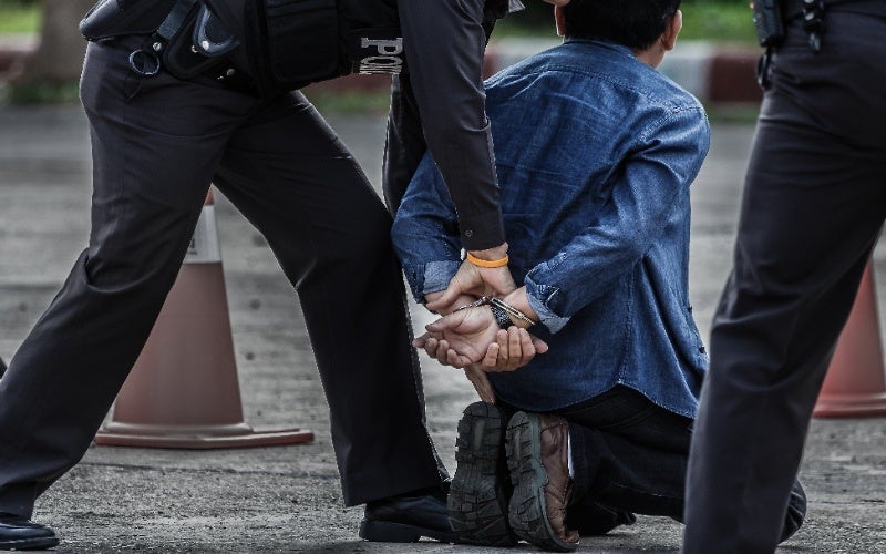 A handcuffed person in a blue denim shirt kneels on the ground as a police officer stands over them.