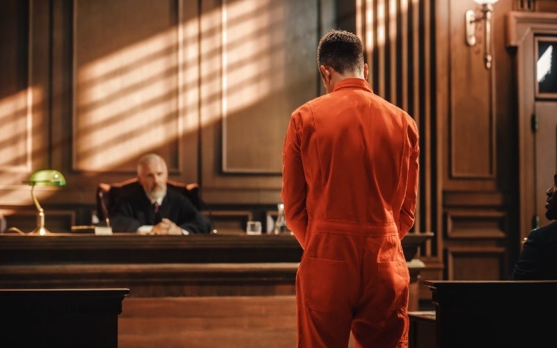 A person in orange jumpsuit stands before a judge in a wood-paneled courtroom.
