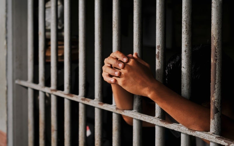 A person's hand clutches against dark metal bars, suggesting confinement and distress.