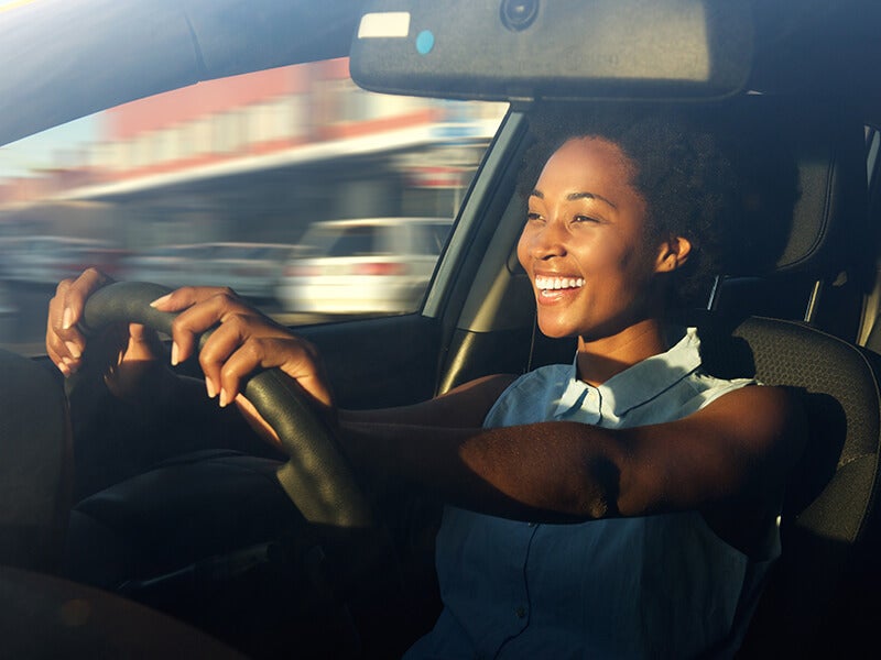 Smiling young Black woman driving, hands on the steering wheel, sun shining on her face. Blurred background implies motion.