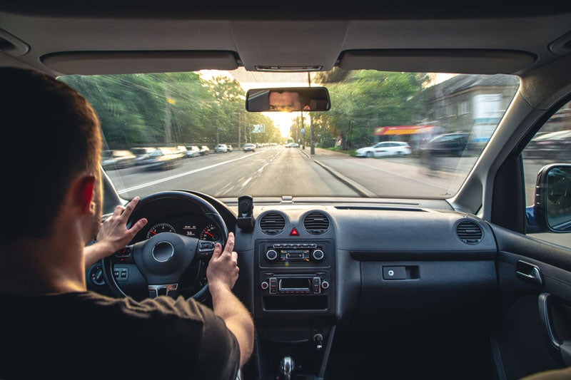 View from a car's interior looking out, a driver with hands on the steering wheel, driving on a sunny street.