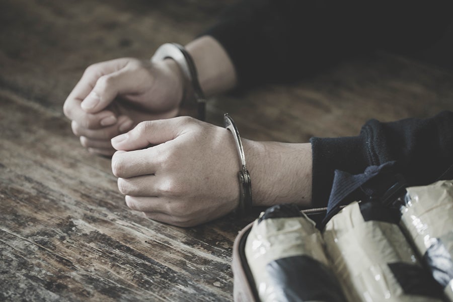 A person's cuffed hands rest on a wooden table next to several tape-wrapped packages of drugs in a container.