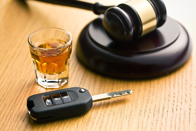 A gavel, glass of whiskey, and car keys rest on a wooden surface, suggesting legal consequences.