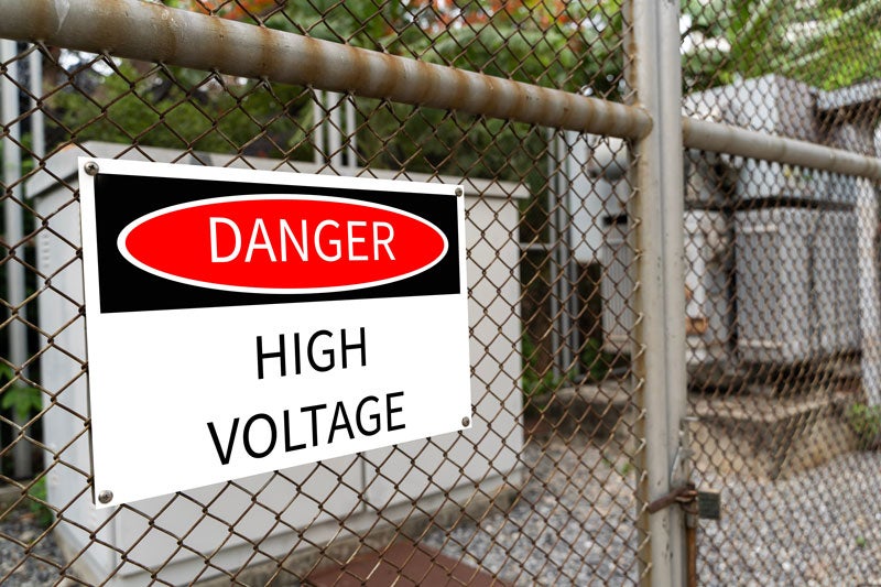 A white sign with DANGER HIGH VOLTAGE in black and red, attached to an old chain-link fence.