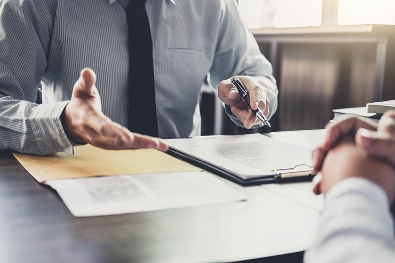 A man in a shirt and tie explains something, gesturing with his hands over documents on a desk.