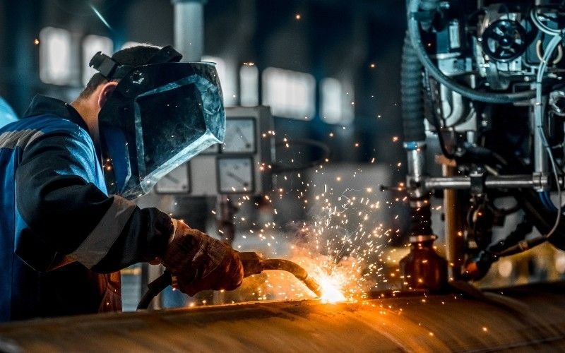 A person in a welding mask and gloves operating a welding torch that emits bright sparks onto a metal pipe.