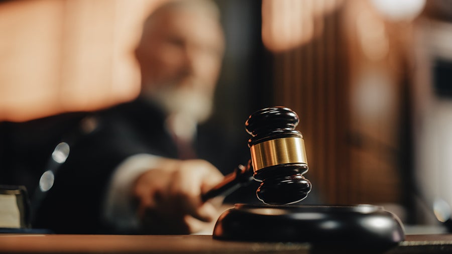 A judge in a blurred background preparing to strike a wooden gavel on a sounding block.