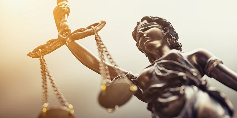 A close-up, low-angle shot of a bronze statue of Lady Justice with golden light reflecting off her scales.