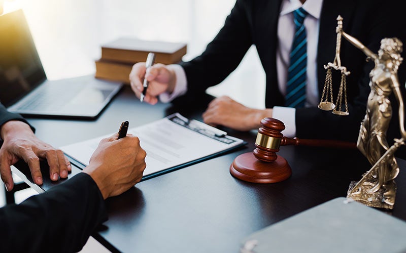 Two people negotiating legal documents at a desk with a gavel and Lady Justice statue.
