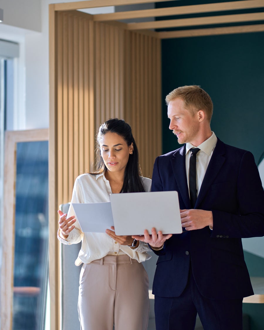 Two business professionals, a woman and a man, discussing work while holding a document and a laptop.