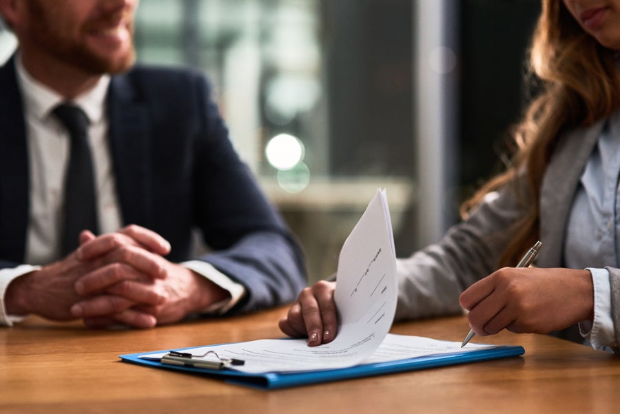 Hands of two people, one signing a document on a clipboard and another watching, representing a business meeting.