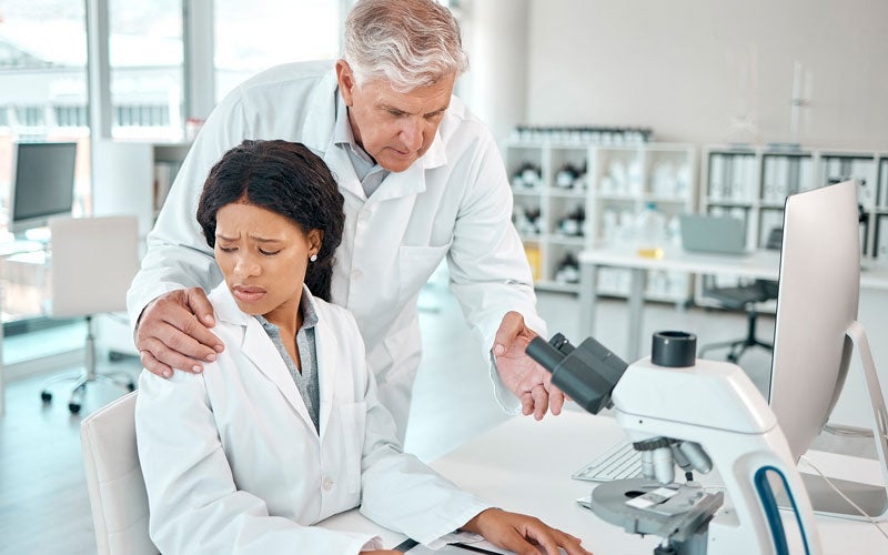 A woman in a lab while a white man places a hand on her shoulder.