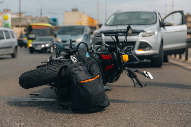 A motorcycle lies on its side in the road after an accident, with a silver SUV and other cars in the background.