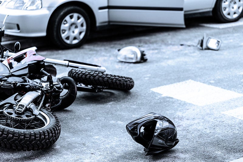 A crashed motorcycle lies on the pavement next to a car with an open door and detached parts.