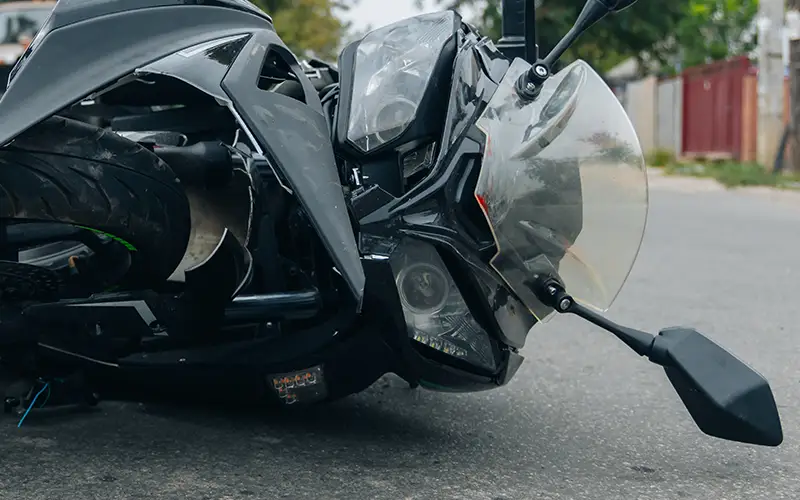 A damaged black motorcycle lies on its side on asphalt, showing a broken headlight and a detached tire.