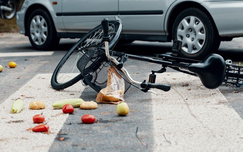 A bicycle and spilled groceries lay on a crosswalk with a silver car visible in the background, suggesting an accident.