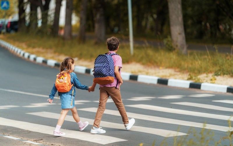 Two children with backpacks, a boy and a girl, holding hands while crossing the street at a crosswalk.