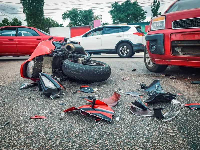 A red motorcycle lies damaged on the pavement surrounded by debris near parked cars.