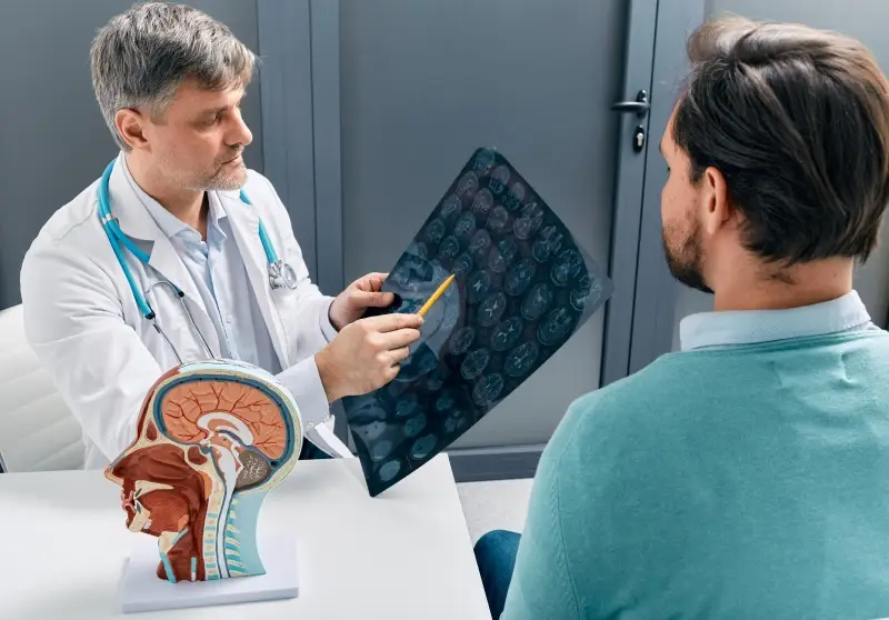 A doctor points to a brain MRI with a pencil while explaining it to a patient in a consultation.