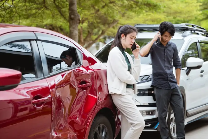 Two people leaning on their damaged cars, talking on phones after an accident. One red, one white car.