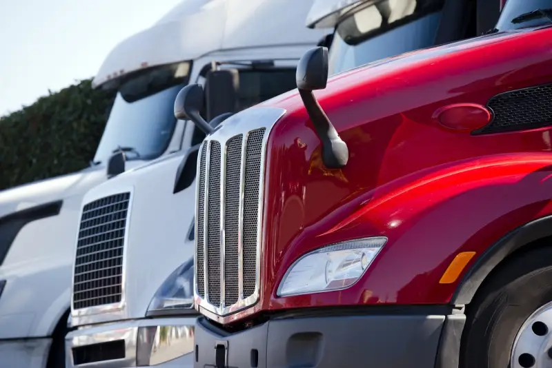 A shiny red semi-truck parked next to a white semi-truck, both seen from the front at an angle.