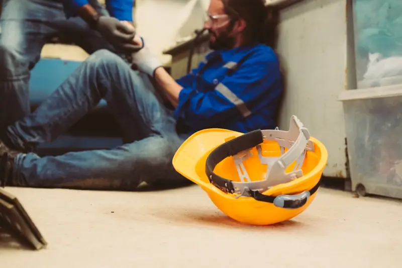 A worker in a blue uniform and safety glasses sits on the floor after an accident, helped by another person, near a fallen yellow hard hat.