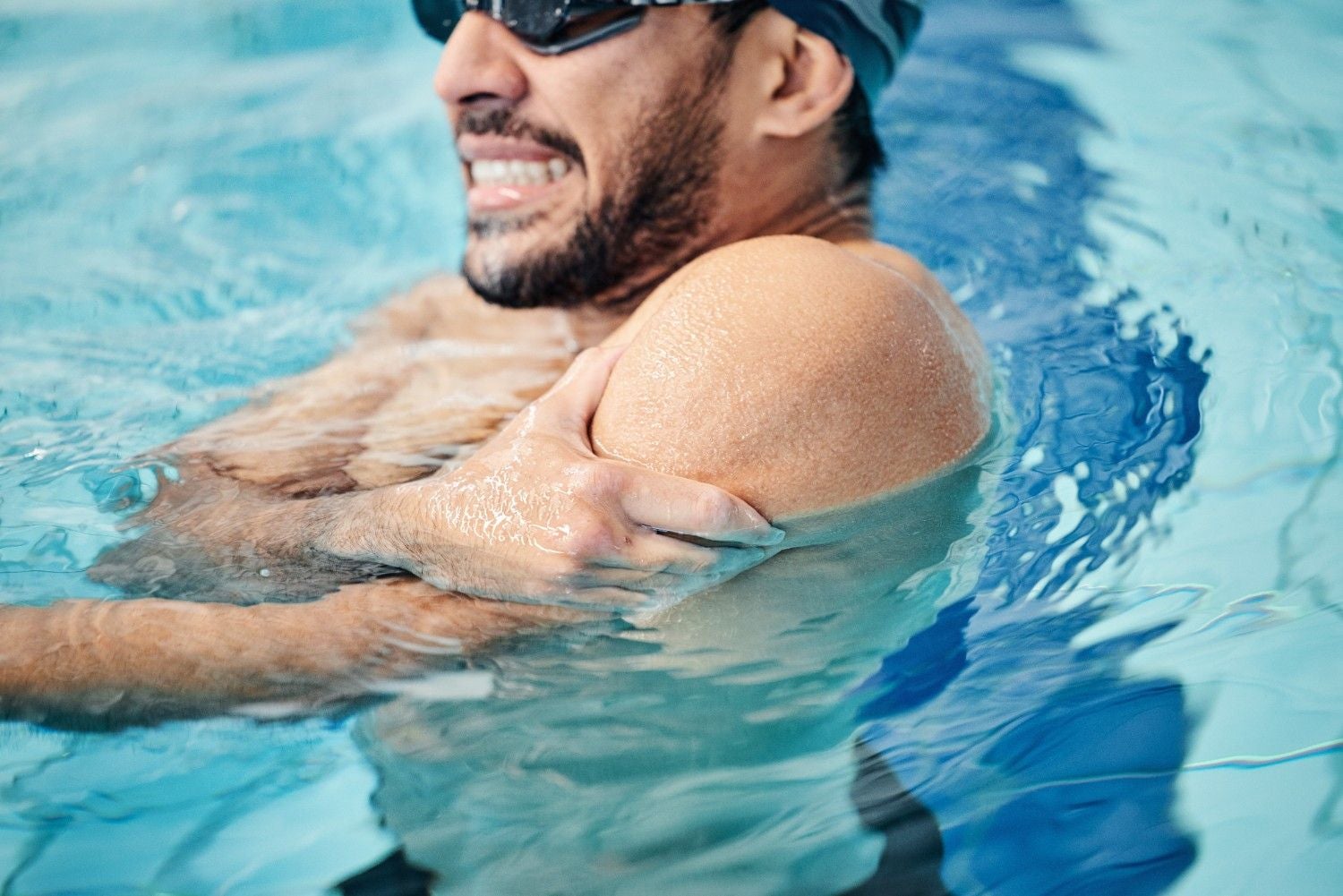 A bearded swimmer in goggles grimaces in pain, clutching his left shoulder with his right hand in a blue pool.