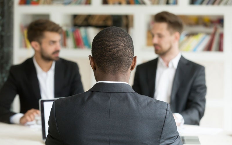 Back of a Black man's head and shoulders, facing two white men in suits sitting across a table.