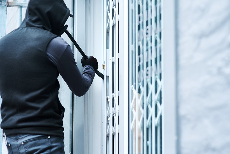 A person in a black hoodie and gloves uses a crowbar to pry open a locked metal exterior gate.